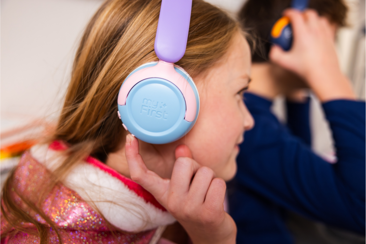A young girl wearing her CareBuds MAX over-ear headphones, adjusting the volume, while her brother is in the background doing the same thing but in softer focus.