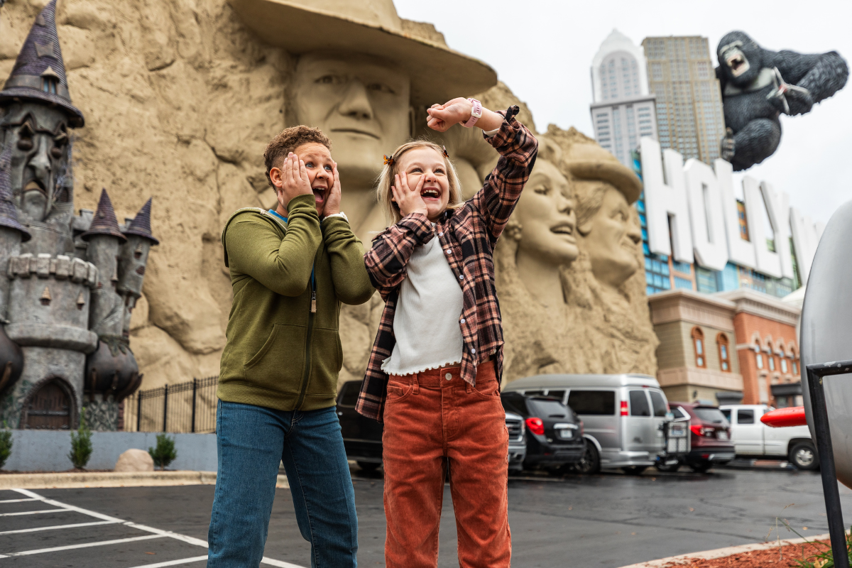 Two kids taking silly pictures with the Fone R2 Kids GPS Smartwatch at a theme park outdoors in a parking lot.