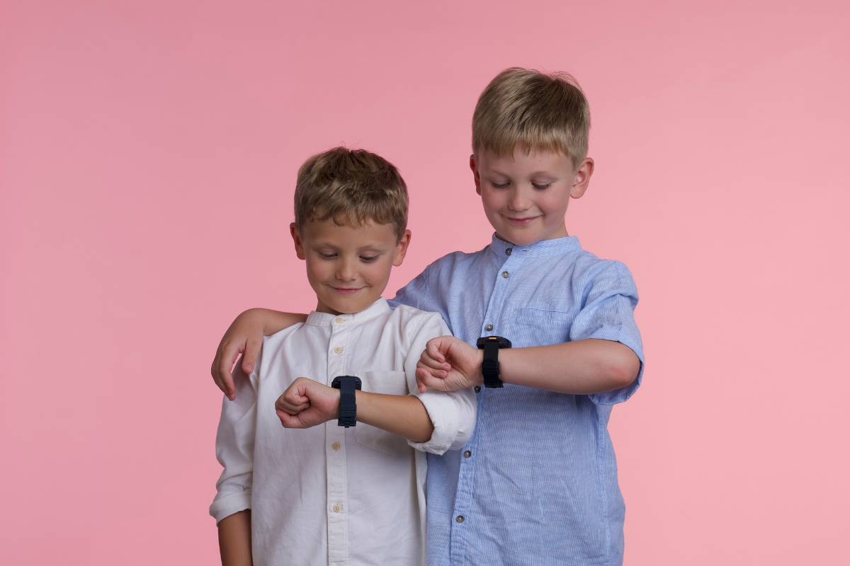 Two young kids looking down at the kids smartwatches on their wrists against a pink background.