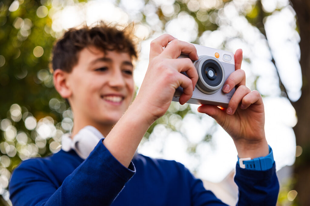 A tween boy using the myFirst Insta Lux camera outside. He's wearing a blue sweater and the camera is white and gray.