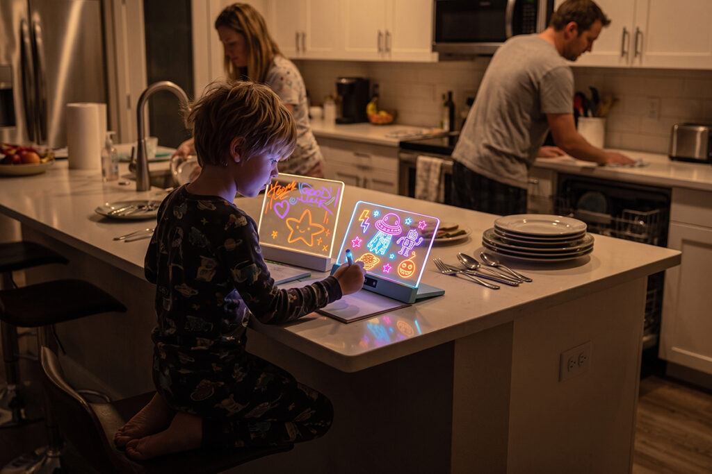 A boy using a glowing Sketch Board Clear in his kitchen while his parents clean up in the background
